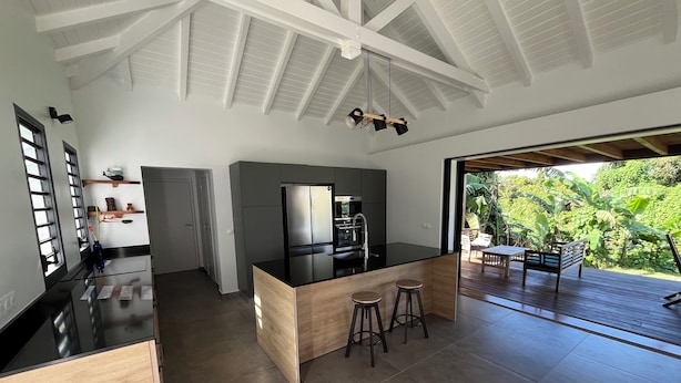 A modern kitchen with a central island, a dining area with a view of lush greenery, and a spacious open ceiling. - Villa le Paradis Secret: Sea view, river access. (Goyave)