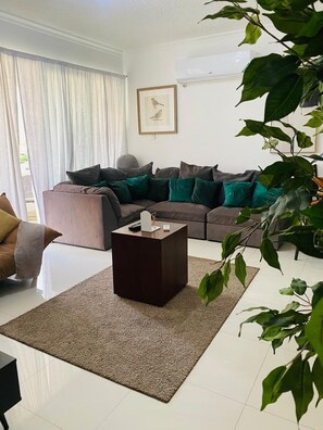 Ein Wohnzimmer mit einer Eckcouch, einem Couchtisch und einem großen Fenster mit Vorhängen.