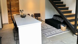 Une cuisine moderne avec un îlot central blanc, des chaises noires et un canapé noir. Un escalier en bois mène à une mezzanine.