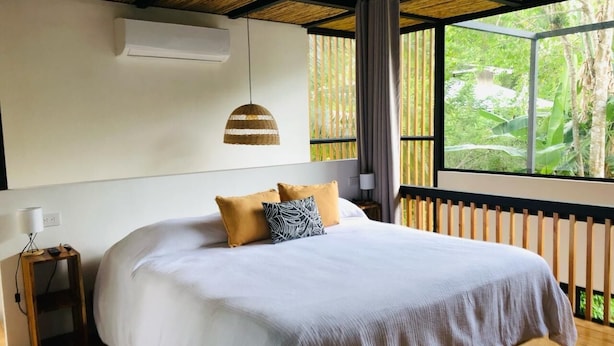 Une chambre avec un grand lit, un luminaire suspendu, une table de chevet et une vue sur la verdure par la fenêtre.
