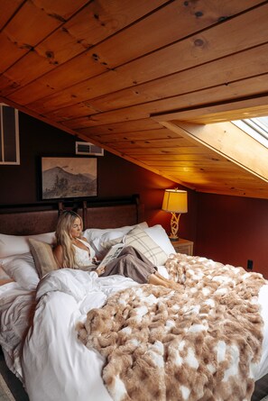 A bedroom with a wooden ceiling, a bed with white bedding, a fluffy brown blanket, a nightstand with a lamp, and a framed picture on the wall.