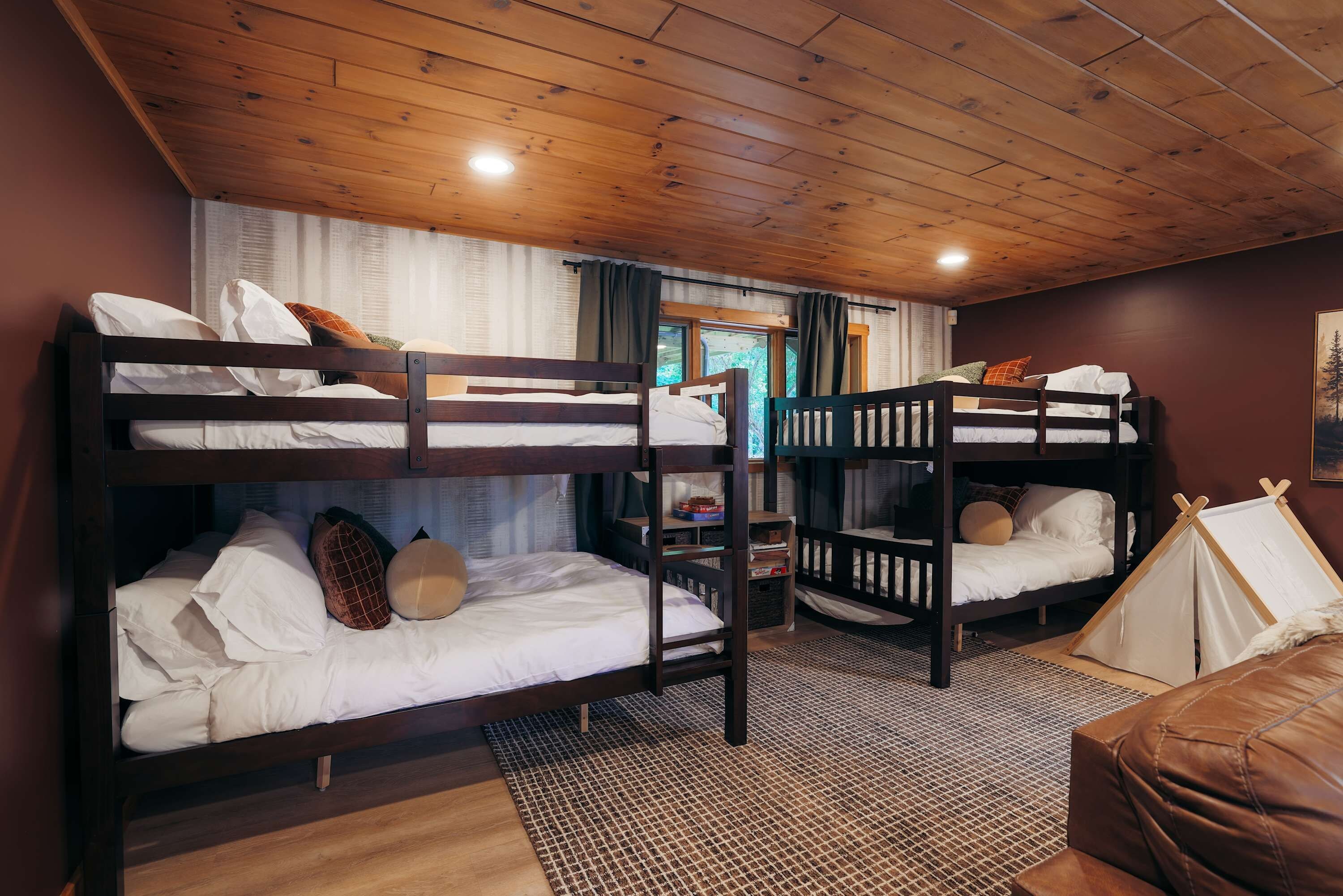 A bunk bed room with a wooden ceiling, a window with curtains, and a tent set up against the wall.