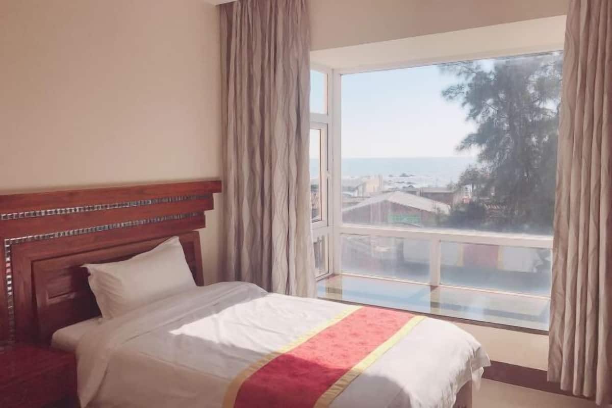 Ein Hotelzimmer mit einem großen Bett, einem Holzkopfende, einem Fenster mit Meerblick und Vorhängen.