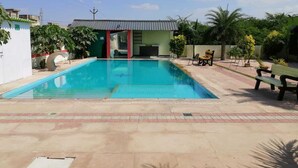 Una piscina con solárium de baldosas, una zona de estar cubierta y un pequeño edificio con puerta roja.