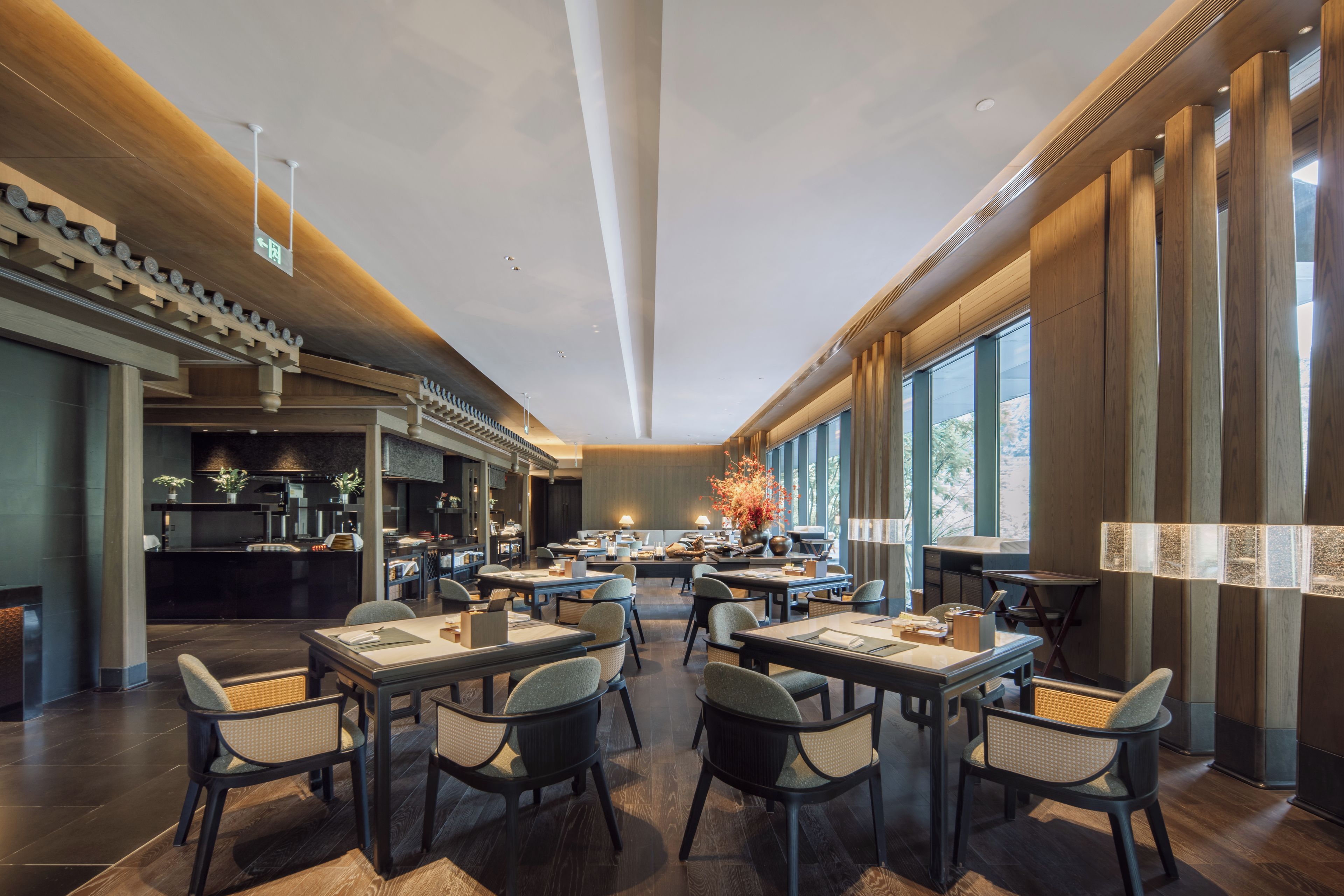 Un restaurante moderno con zona de comedor central, cocina al fondo y grandes ventanales a lo largo de una pared.