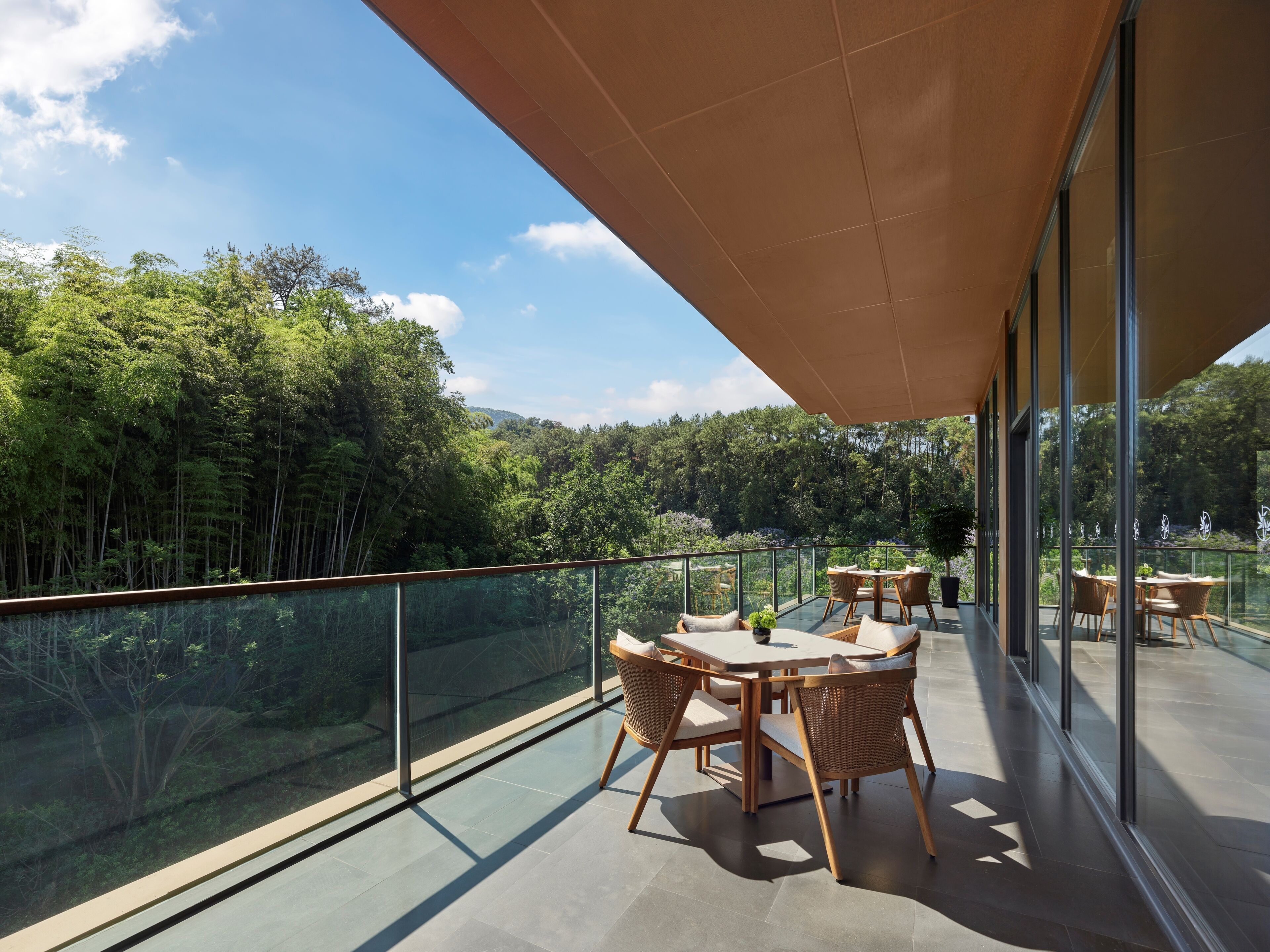 A balcony with glass railings, a table with chairs, and a view of a forested area.