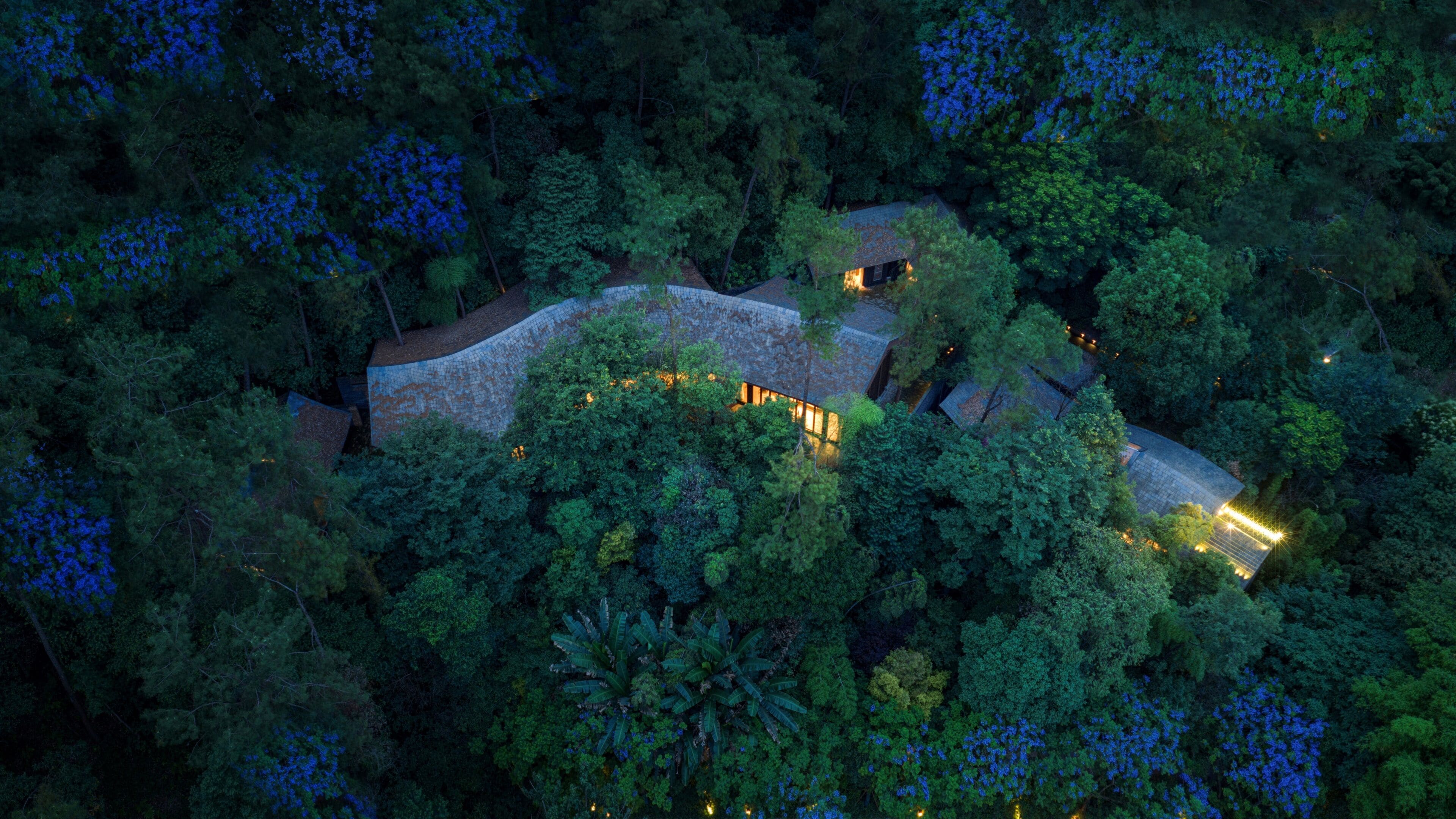 An aerial view of a secluded, tree-covered estate with multiple buildings, illuminated pathways, and a central structure with a distinctive roof.