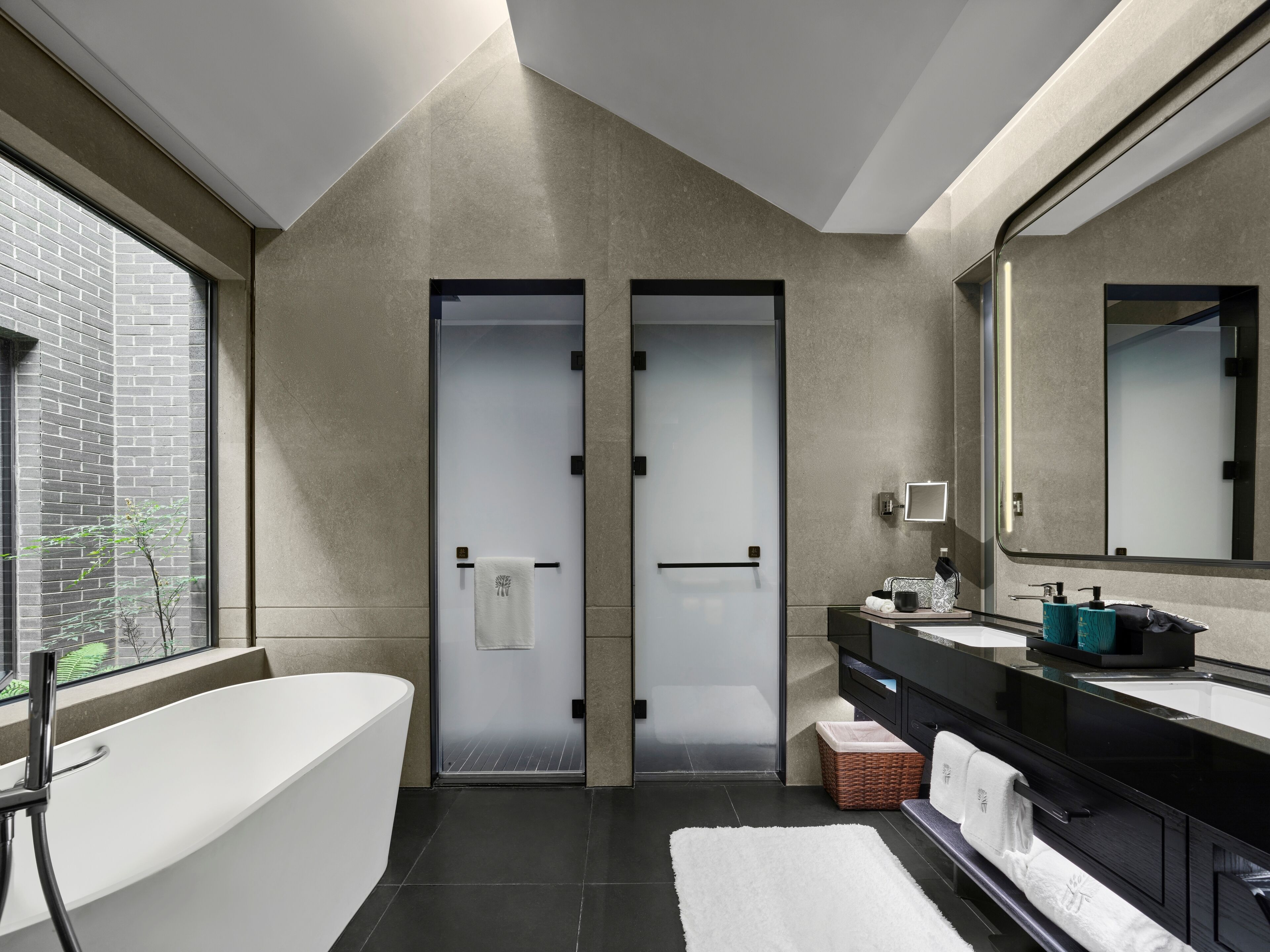 A modern bathroom with a freestanding bathtub, a large mirror, and a window with a view of a brick wall.