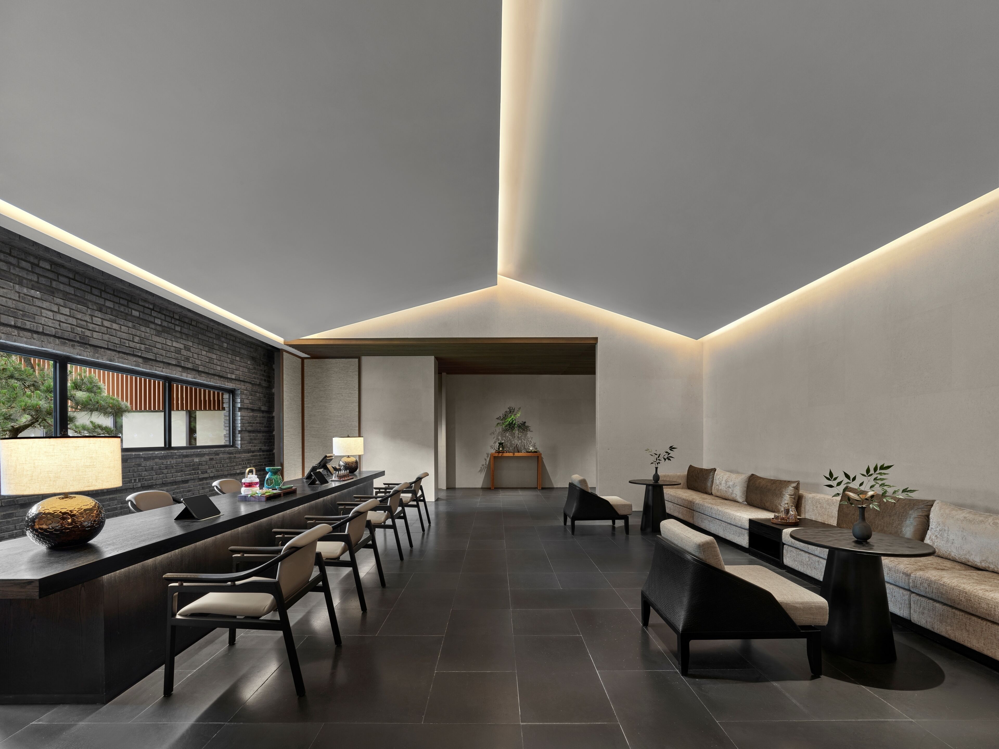 A modern interior with a long reception desk, seating area, and a view of a tree outside.