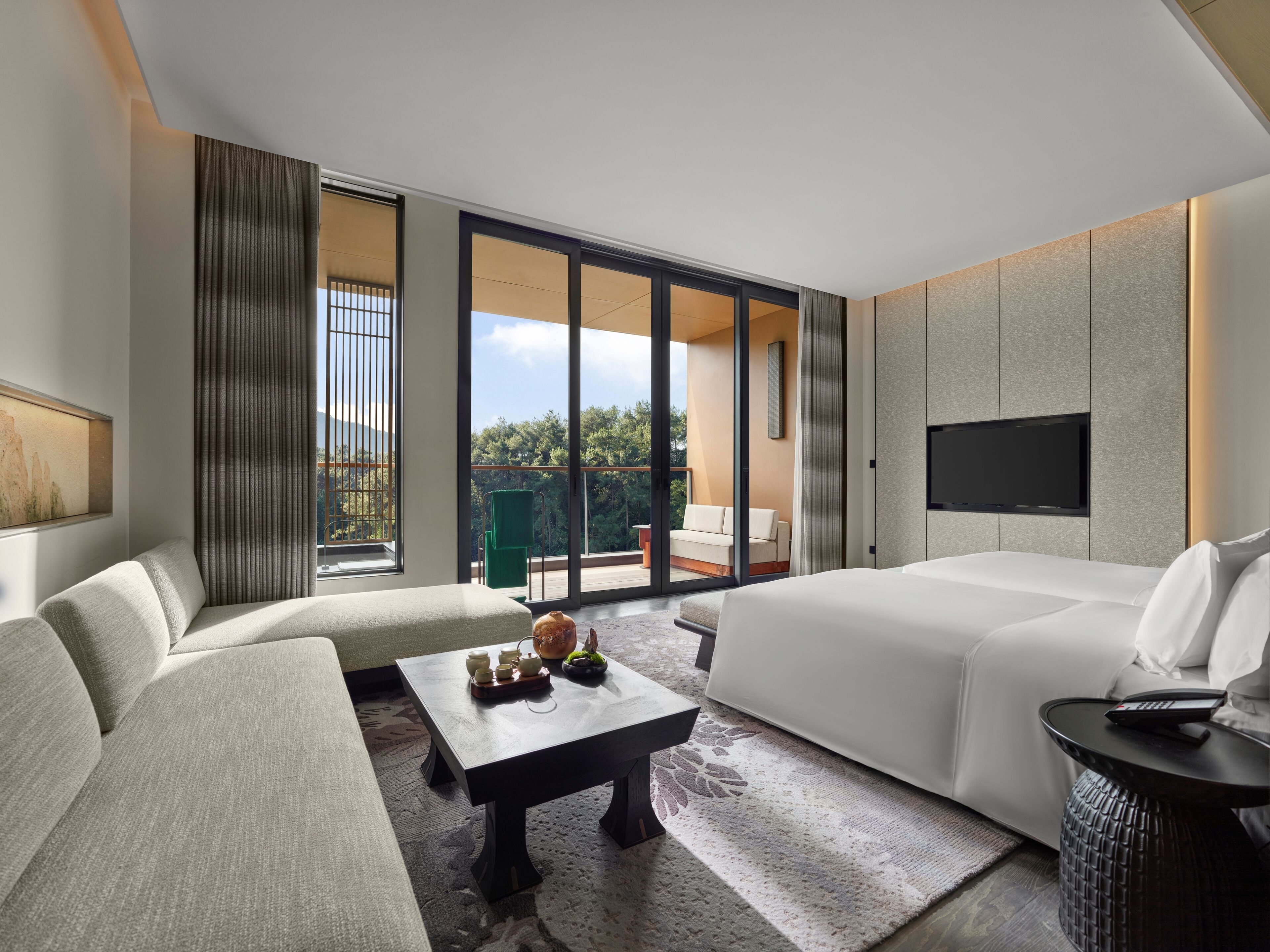 Una habitación de hotel moderna con sofá, cama, mesa de centro, televisor y balcón con vistas a árboles.
