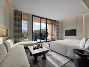 Une chambre d’hôtel moderne équipée d’un canapé, d’un lit, d’une table basse, d’une télévision et d’un balcon avec vue sur les arbres.