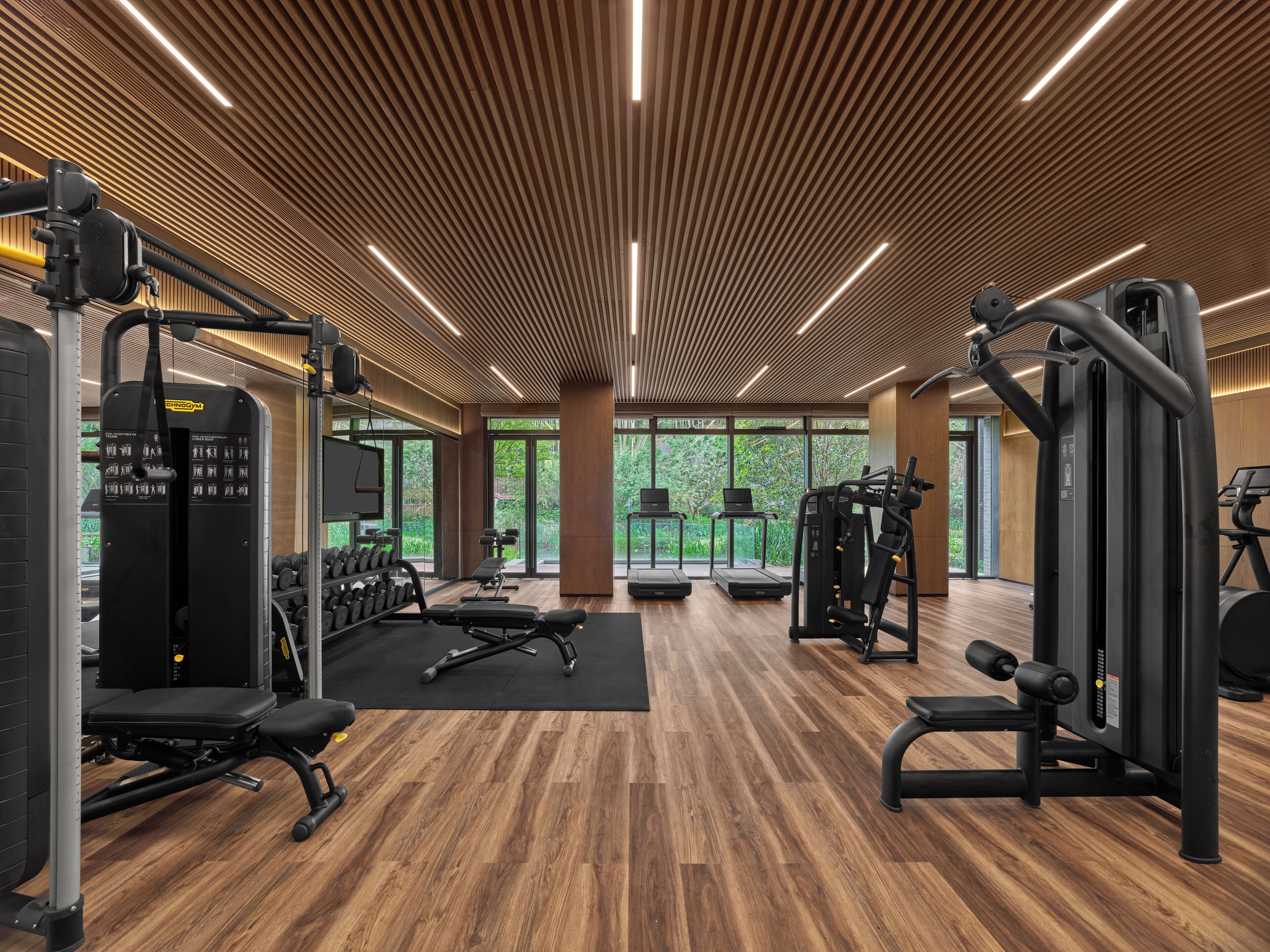 Un gimnasio moderno con suelo de madera, equipamiento de ejercicio y grandes ventanales que dan a zonas verdes.