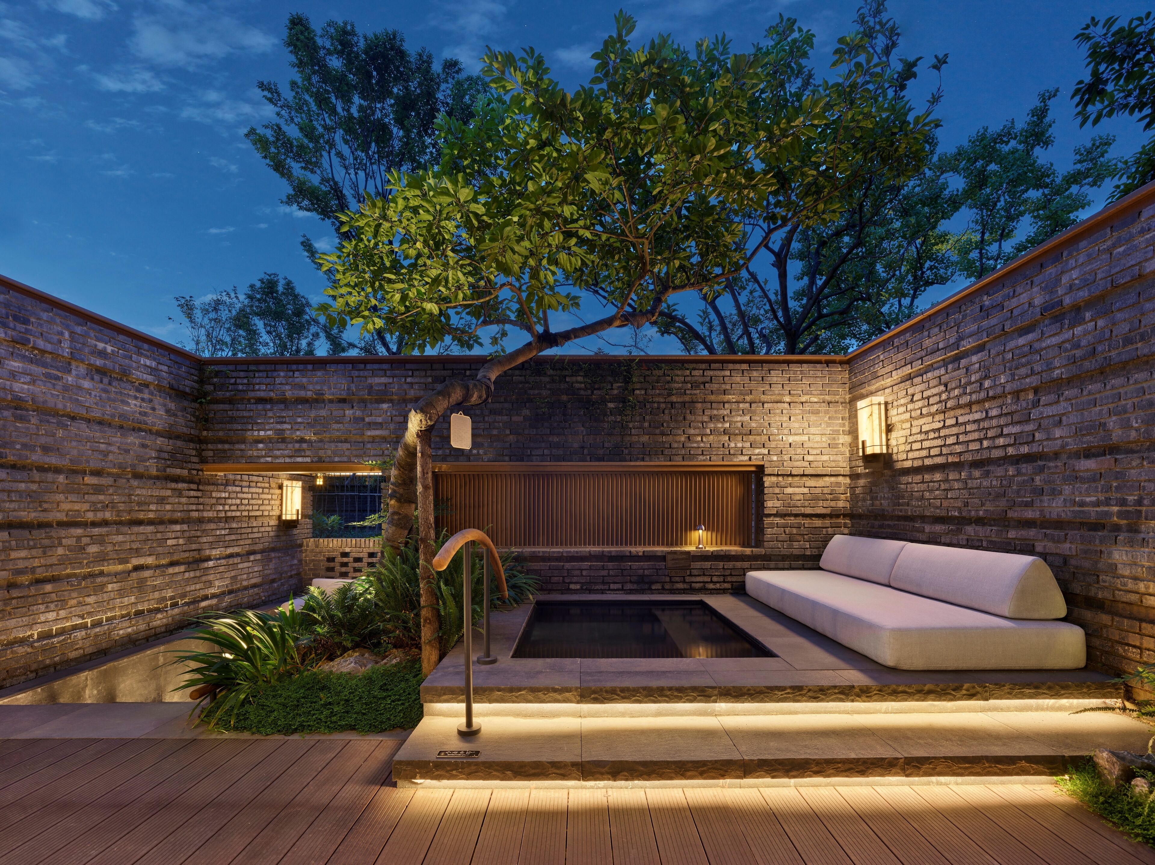 Eine moderne Außenpatio mit Backsteinmauer, einer Holzbank, einem kleinen Pool und einem Baum.