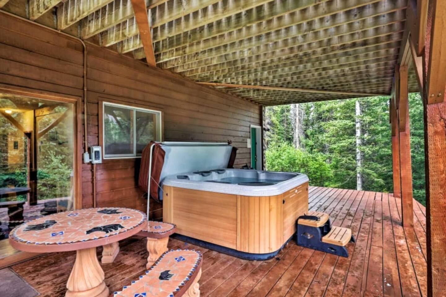 Covered wooden deck featuring a hot tub and an outdoor tiled table overlooking the woods.