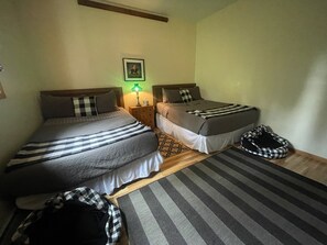 Ein Zimmer mit zwei Einzelbetten, einem Nachttisch mit Lampe, einem karierten Haustierbett und einem gerahmten Bild an der Wand.