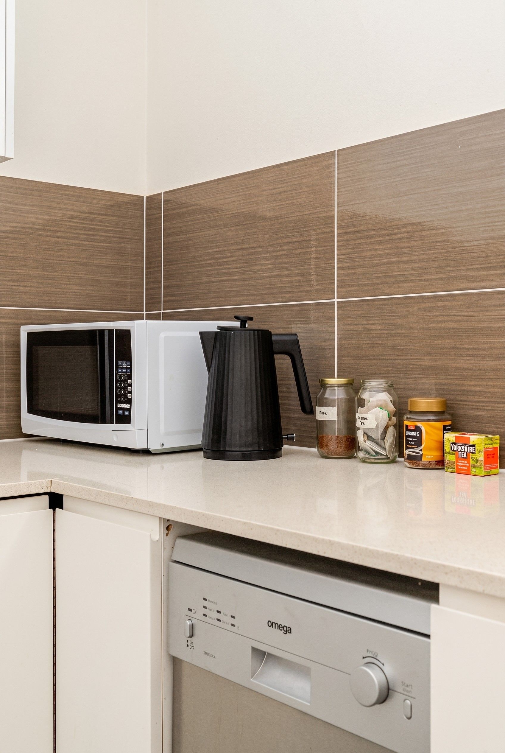 Een keuken met een magnetron, een zwarte elektrische waterkoker en theekruiden in potten op het aanrecht.