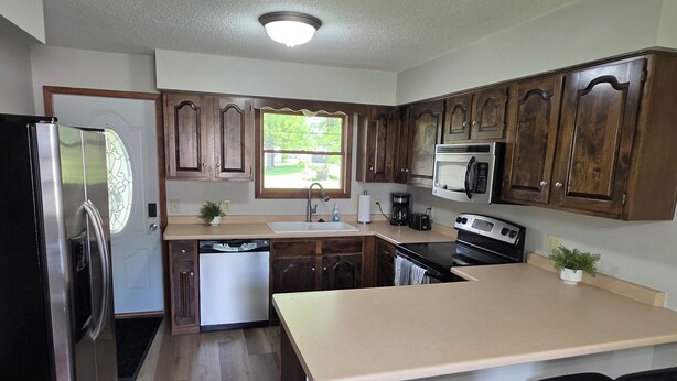 A kitchen with wooden cabinets, a stainless steel refrigerator, a dishwasher, a microwave, an oven, a sink, and a window with a view of greenery.