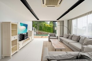 A modern living room with a large TV, a sofa, a coffee table, and a view of a pool and greenery outside. - Tranquil 3-Storey Villa with Private Pool (Koh Samui)