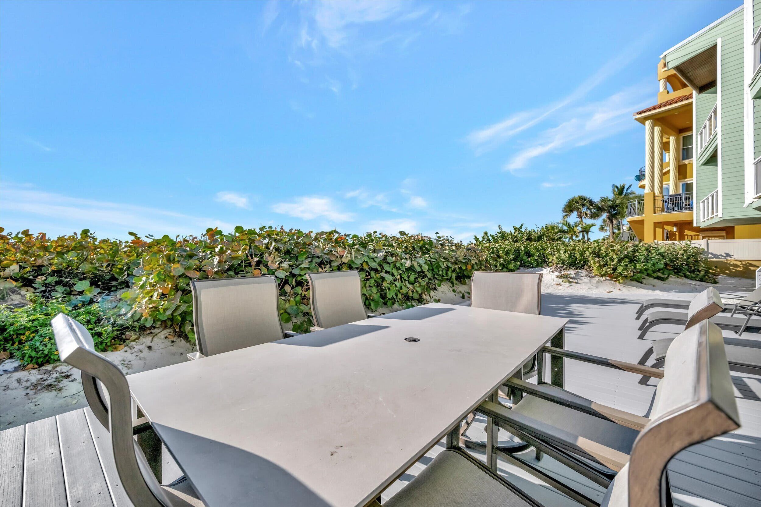 An outdoor patio with a table and chairs, surrounded by a hedge and a building with balconies.