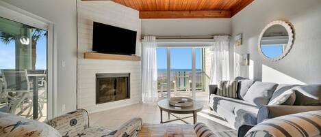 A spacious living room with a wooden ceiling, a fireplace, a TV mounted on the wall, and a large window offering a view of the outdoors.