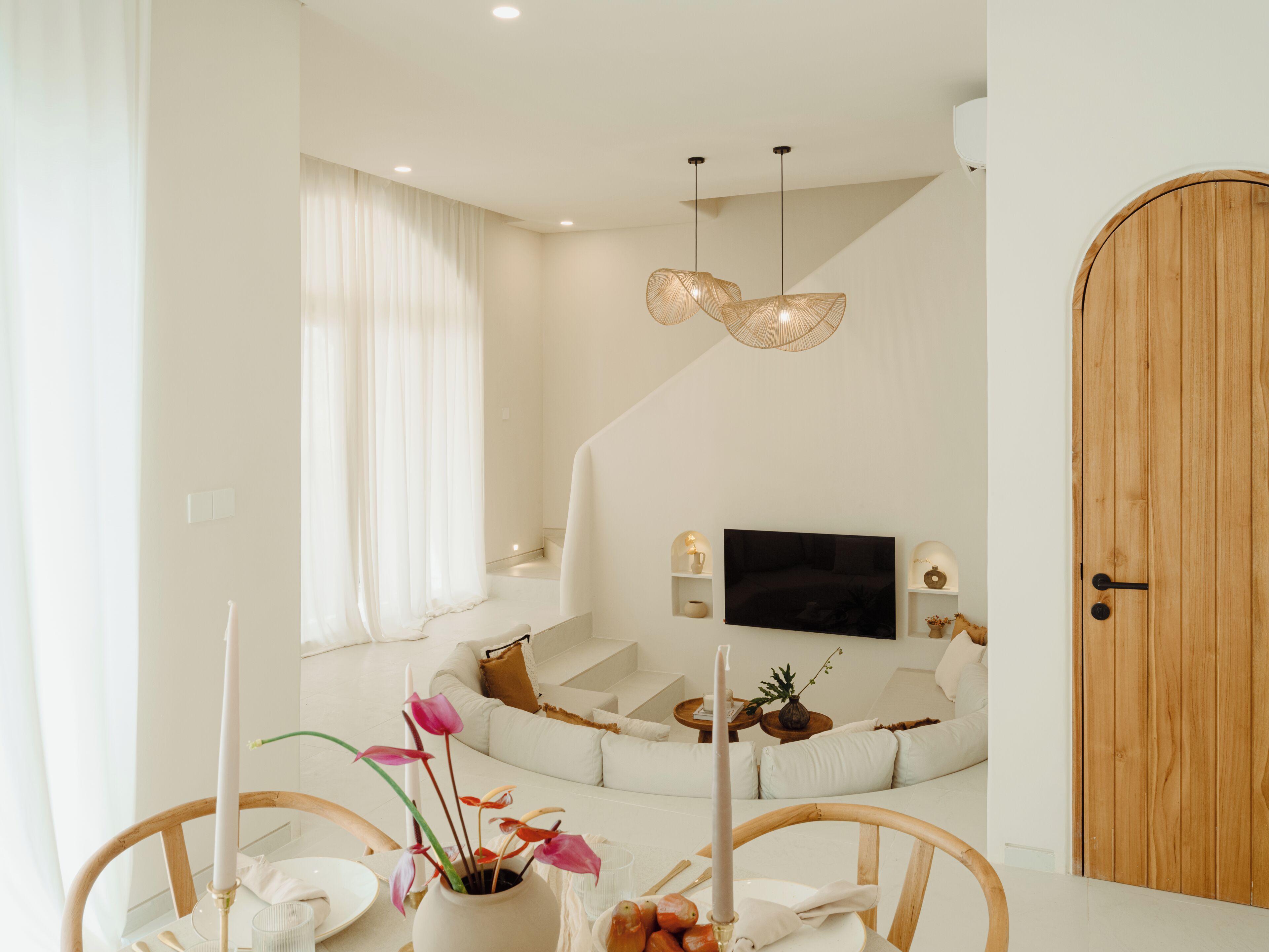 A modern living room with a sofa, a dining table with chairs, a TV mounted on the wall, and a wooden door.