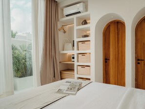 Une chambre à coucher comprenant un lit, une fenêtre avec des rideaux, un appareil de climatisation, une porte en bois et des étagères intégrées avec des paniers de rangement.