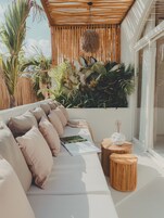 A sunlit outdoor seating area with a sofa, cushions, a small table, and a hanging light fixture.