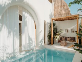 A poolside area with a clear blue pool, a covered seating area with cushions, and a wall with arched niches.