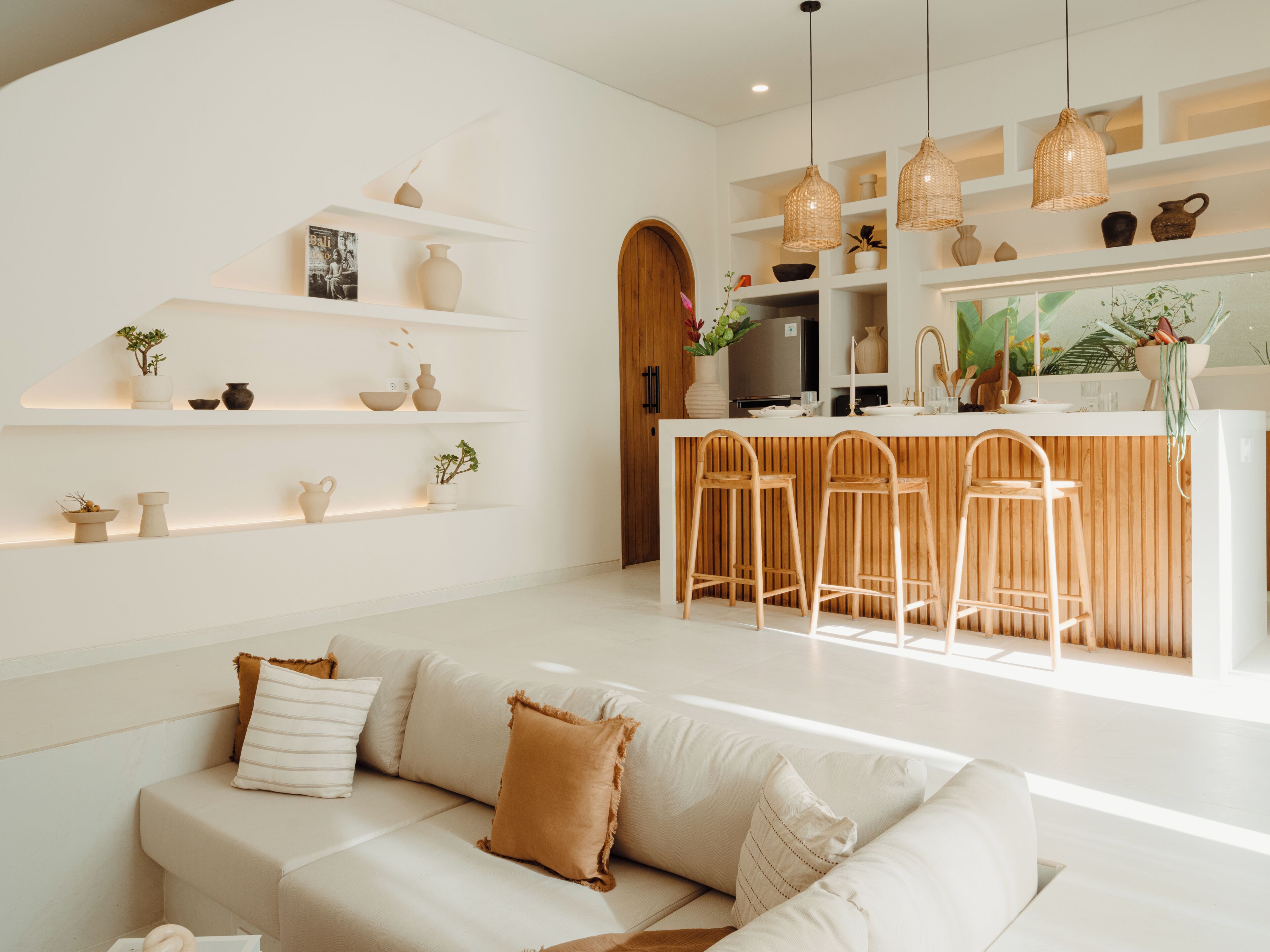 A modern living room with a white sofa, a kitchen area with a bar, and shelves with decorative items.