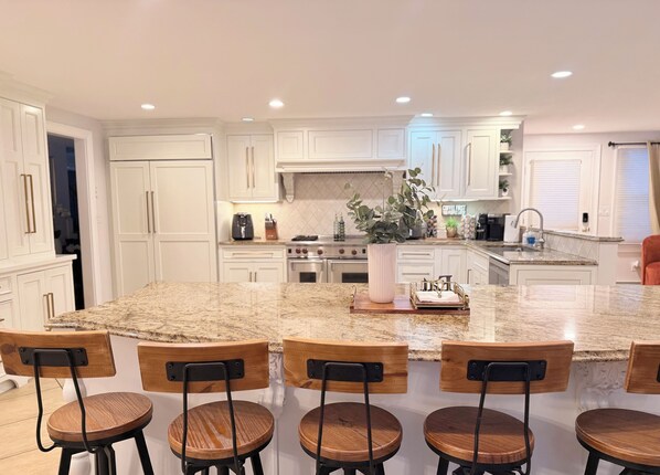 A modern kitchen with white cabinetry, a large island with bar stools, and a central island with a plant. - Beautiful 6-bedroom house with Outdoor patio and heated pool in Plymouth, Ma (Plymouth)