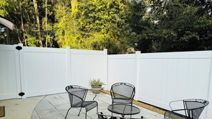A patio with four metal chairs and a round table, surrounded by a white fence and trees.