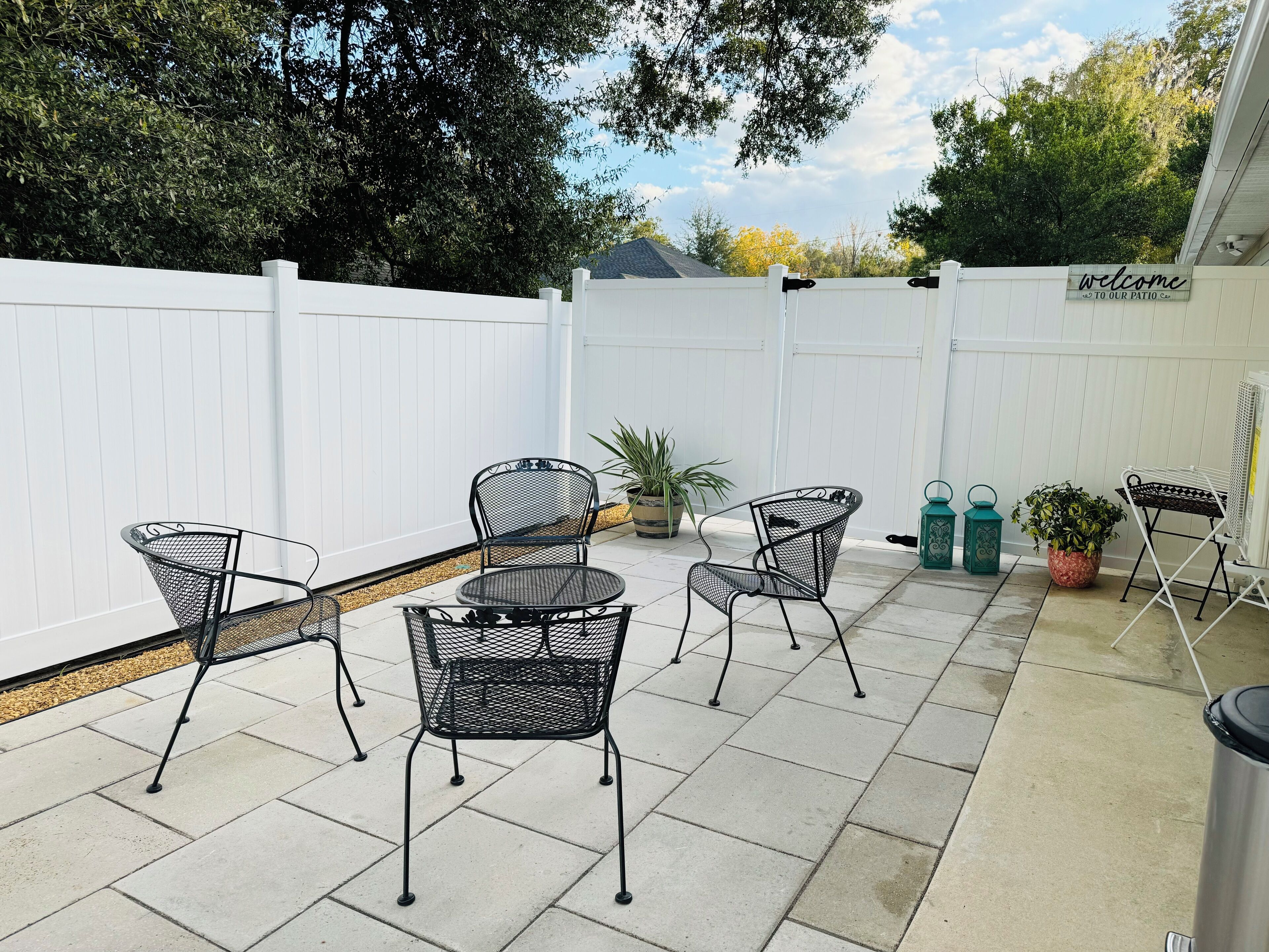 A patio with four metal chairs and a table, surrounded by a white fence, with potted plants and a sign that reads "Welcome to Our Patio."