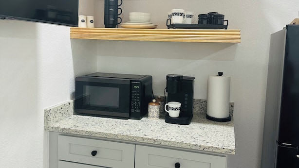 A compact kitchen with a microwave, coffee maker, and a coffee-themed sign on the shelf.