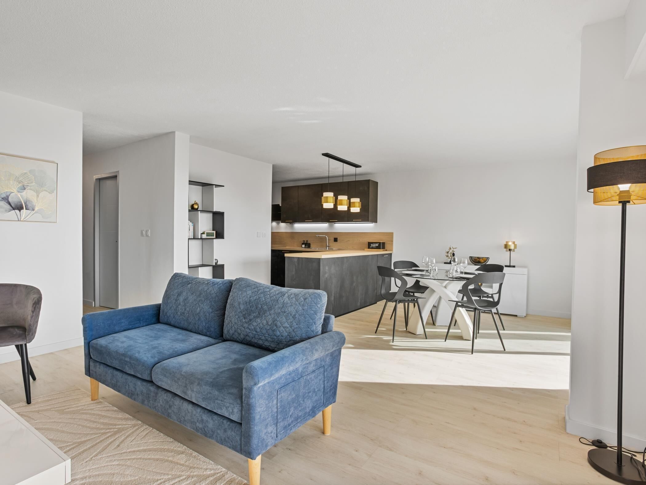 A modern living room with a blue sofa, a dining area with a table and chairs, and a kitchen with dark cabinets and pendant lighting.