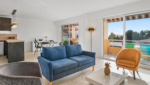 A modern living room with a blue sofa, a grey armchair, and a white coffee table. There is a dining area with a table and chairs, and a kitchen area with a counter and cabinets. The room has large windows and a view of the outside. - 4* - sea view - 2 bedrooms - parking - swimming pool (Saint-Raphaël)