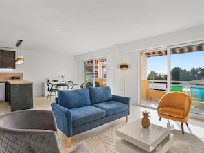 A modern living room with a blue sofa, a grey armchair, and a white coffee table. There is a dining area with a table and chairs, and a kitchen area with a counter and cabinets. The room has large windows and a view of the outside. - 4* - sea view - 2 bedrooms - parking - swimming pool (Saint-Raphaël)