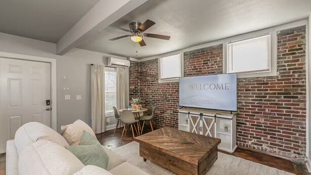 Uma sala de estar moderna com parede de tijolos, mesa de centro de madeira, sofá e TV exibindo “WELCOME”.