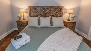 Cama bem arrumada com colcha acolchoada, duas almofadas e um cobertor branco sobre uma cabeceira de madeira. Há duas mesas de cabeceira com abajures e pequenos vasos.