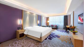 A modern hotel room with a large bed, a desk, a chair, and a small table with a fruit bowl.