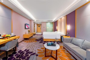 Ein Hotelzimmer mit Bett, Sofa, Schreibtisch mit Stuhl, einem kleinen Tisch und einem durchgehenden Türbogen einsehbarer Badezimmer.