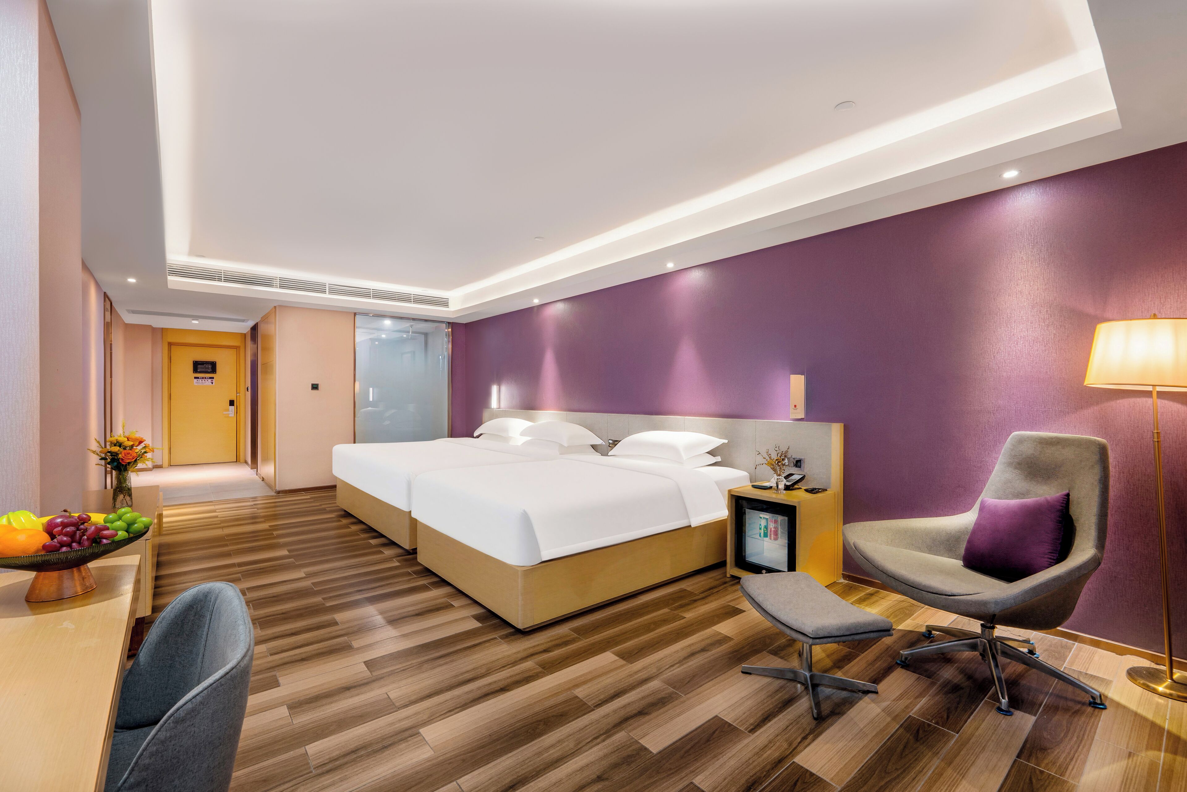 A modern hotel room with a large bed, a desk with a fruit bowl, a chair, and a bedside table with a lamp.