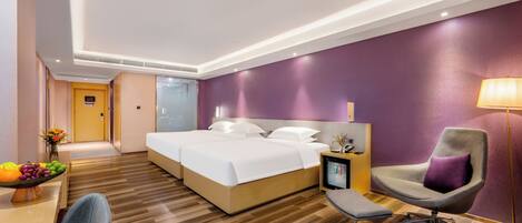A modern hotel room with a large bed, a desk with a fruit bowl, a chair, and a bedside table with a lamp.