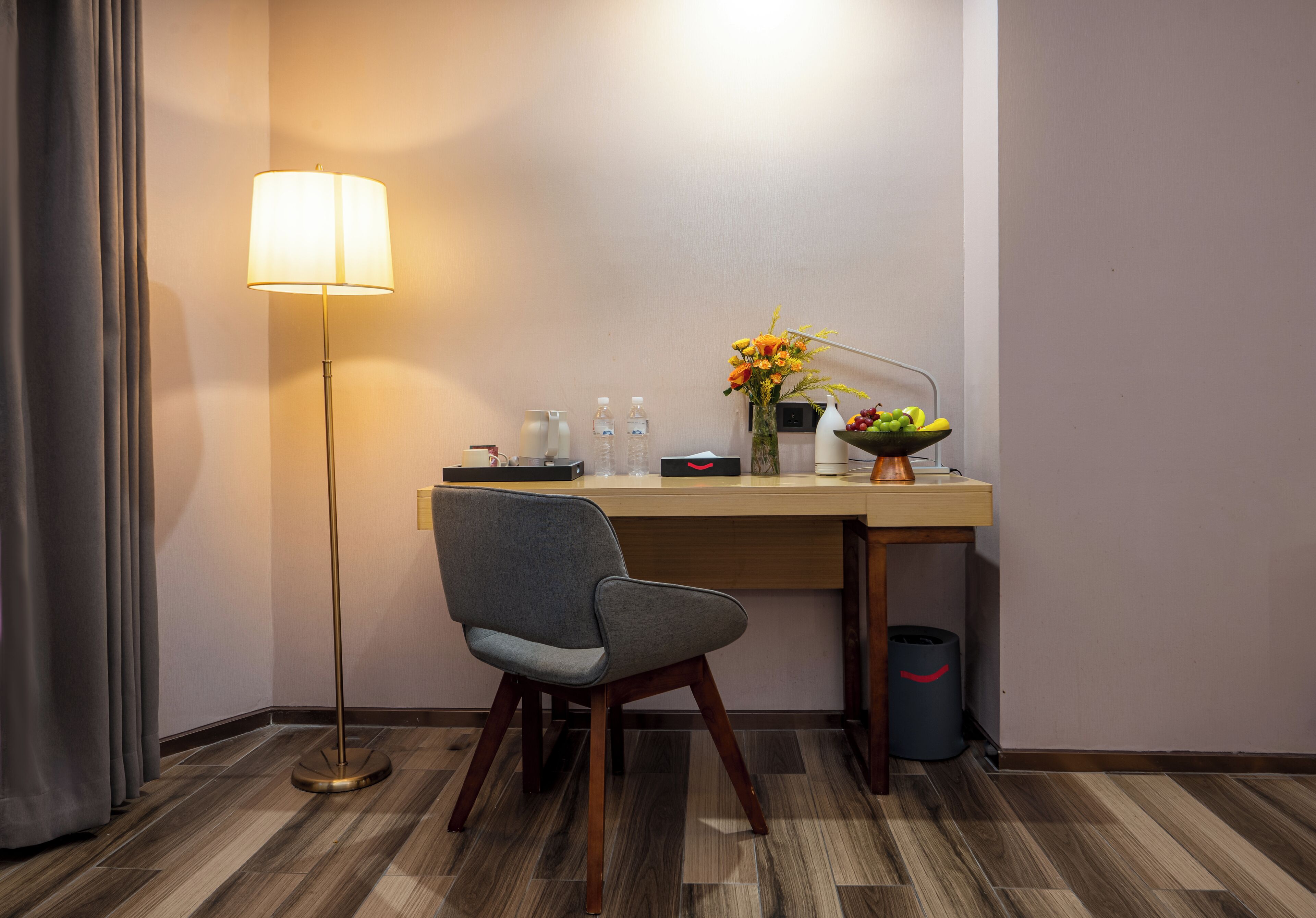 A hotel room with a desk, chair, lamp, flower vase, fruit bowl, and a trash can.