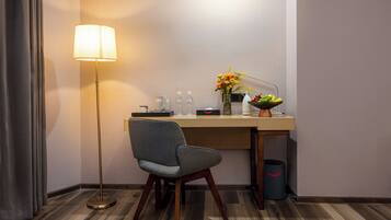 A hotel room with a desk, chair, lamp, flower vase, fruit bowl, and a trash can.
