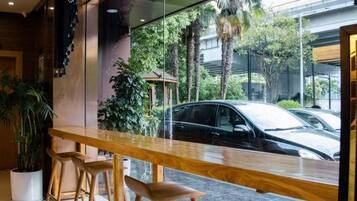 Uma área de bar com balcão de madeira e banquetas, uma janela grande com vista para carros estacionados e vegetação lá fora.