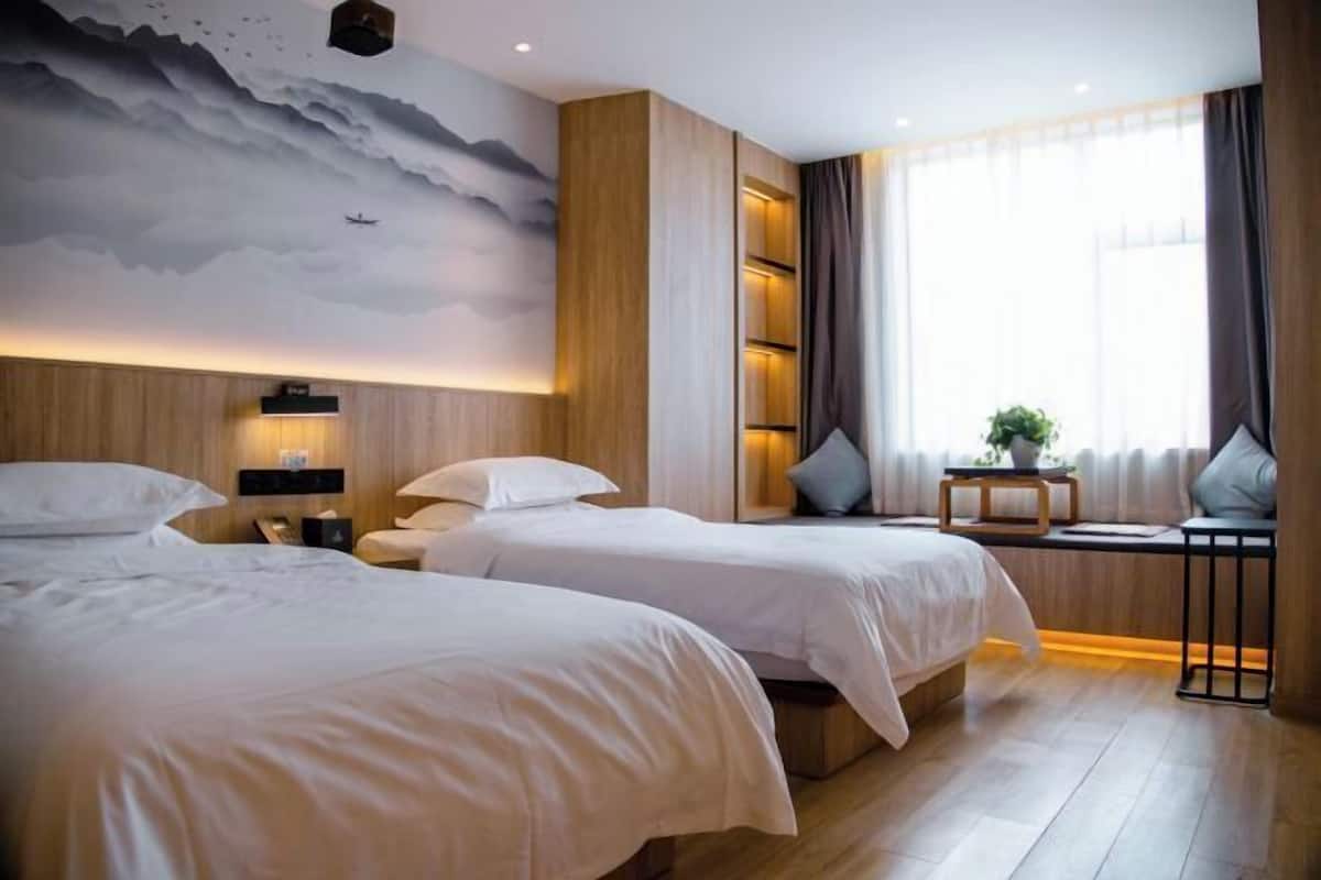 Habitación de hotel con dos camas, un mural en la pared, una ventana con cortinas y una pequeña mesa con una planta.