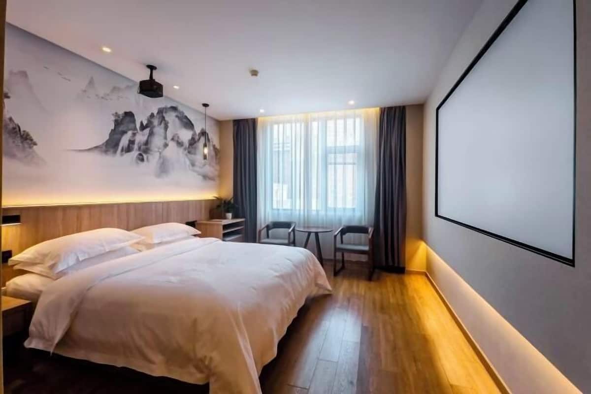 Habitación de hotel con una cama grande, televisor montado en la pared, zona de estar con sillones y una ventana con cortinas.