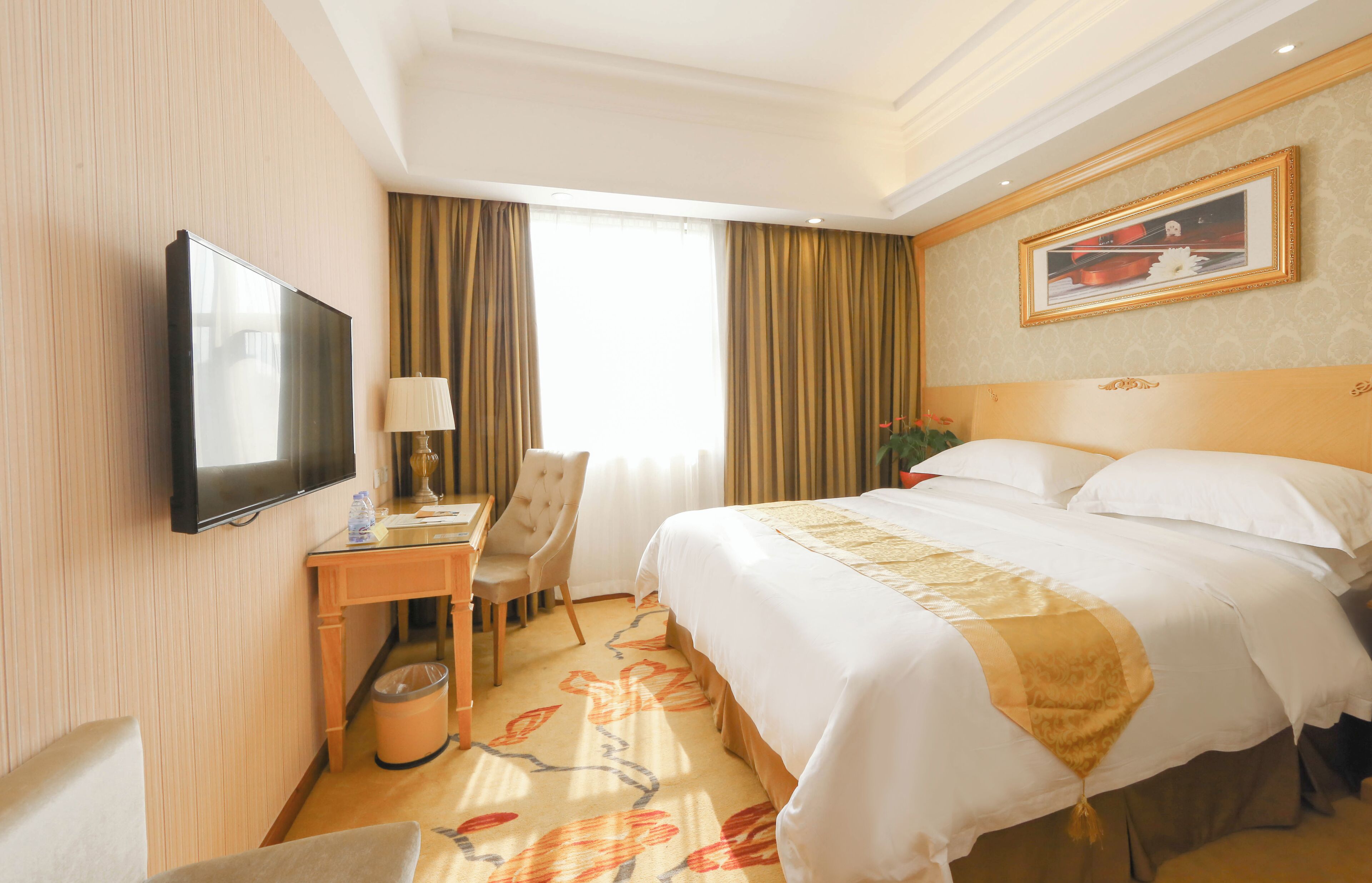 A hotel room with a bed, a desk, a chair, a television, and a painting on the wall.