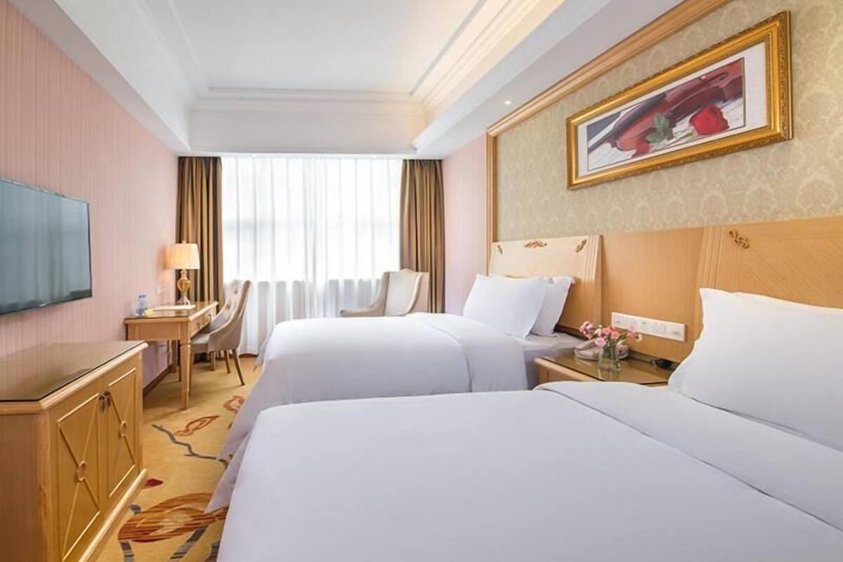 A hotel room with two beds, a desk with a lamp, a television, and a painting on the wall.
