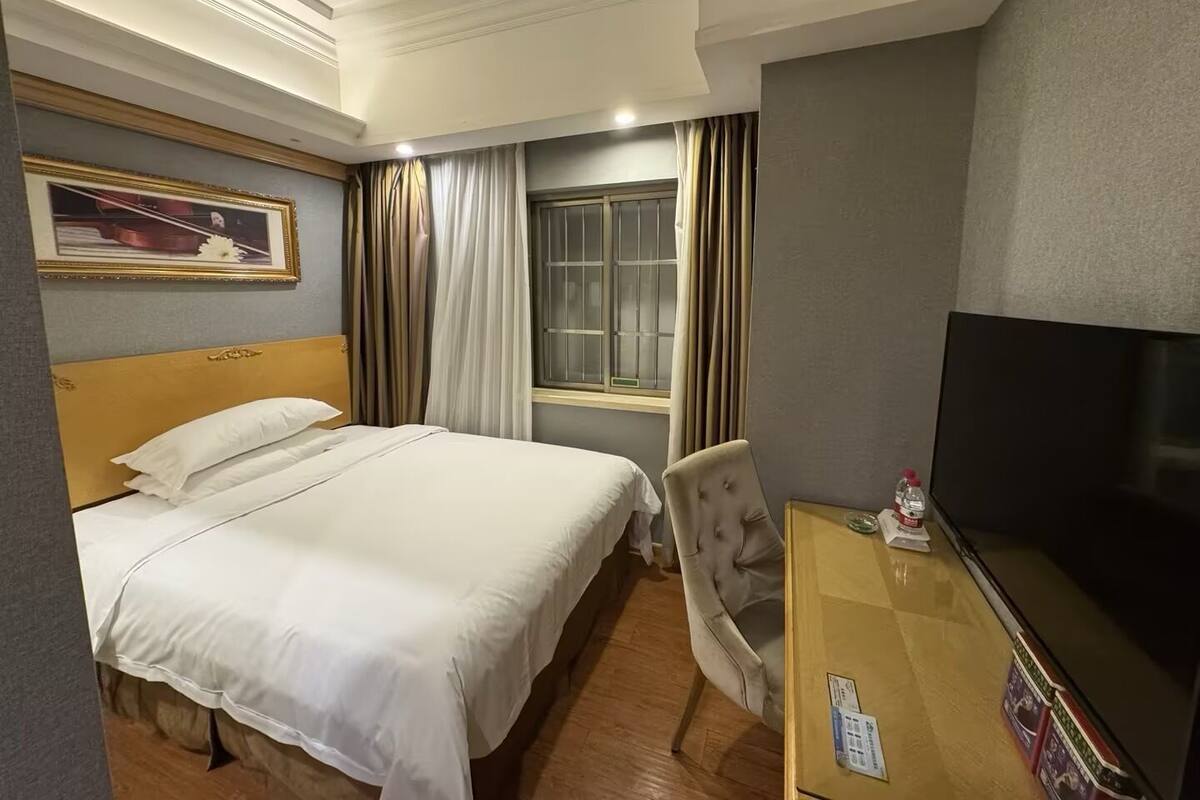 A hotel room with a bed, a TV, a chair, a window with curtains, and a framed picture on the wall.